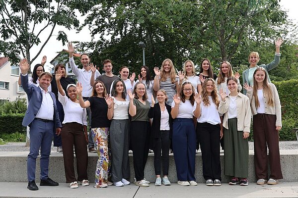 Eine Gruppe von etwa 20 Personen steht im Freien und winkt in die Kamera. Die Personen sind unterschiedlich gekleidet und lächeln. Im Hintergrund sind Bäume und Gebäude zu sehen, die eine freundliche Atmosphäre schaffen.