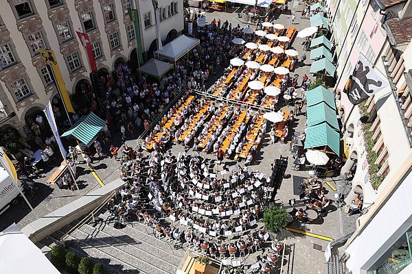 Eine Menschenmenge versammelt sich in einem Platz, umgeben von historischen Gebäuden. Tische befinden sich im Freien, einige sind mit Sonnenschirmen ausgestattet. Im Vordergrund spielt ein großes Orchester. Es ist sonnig und der Platz scheint festlich dekoriert zu sein.
