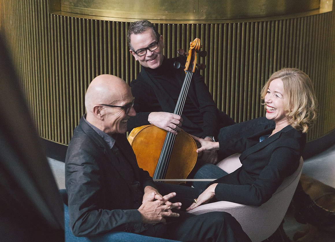 Zwei Männer und eine Frau sitzen lachend zusammen. Einer hält ein Cello in der Hand.