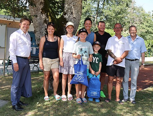 Eine Gruppe von neun Personen steht unter einem Baum im Freien. Sie lächeln für das Foto. Einige halten Taschen, während andere in Freizeitkleidung gekleidet sind. Im Hintergrund sind Bänke und eine Einrichtung sichtbar.