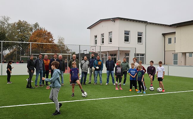 Eine Gruppe von Erwachsenen und Kindern steht auf einem neuen Fußballfeld. Die Kinder halten Fußbälle in den Händen und einige bereiten sich darauf vor, zu spielen. Im Hintergrund sind ein Gebäude und Bäume zu sehen.