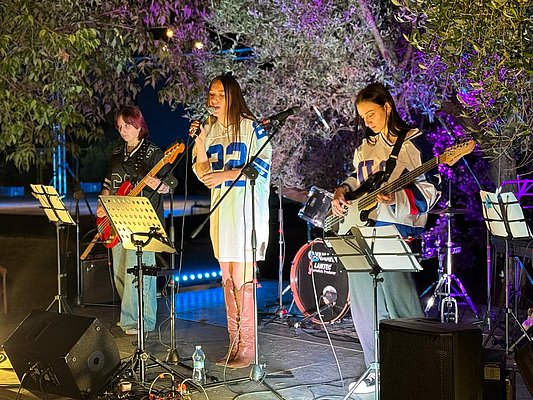Eine Band spielt live auf einer Bühne im Freien. Die Sängerin in einem weißen Kleid tritt vor das Publikum, während zwei Musiker neben ihr an einer E-Gitarre und einer Bassgitarre spielen. Im Hintergrund sind Schlagzeug und Lichter zu sehen.