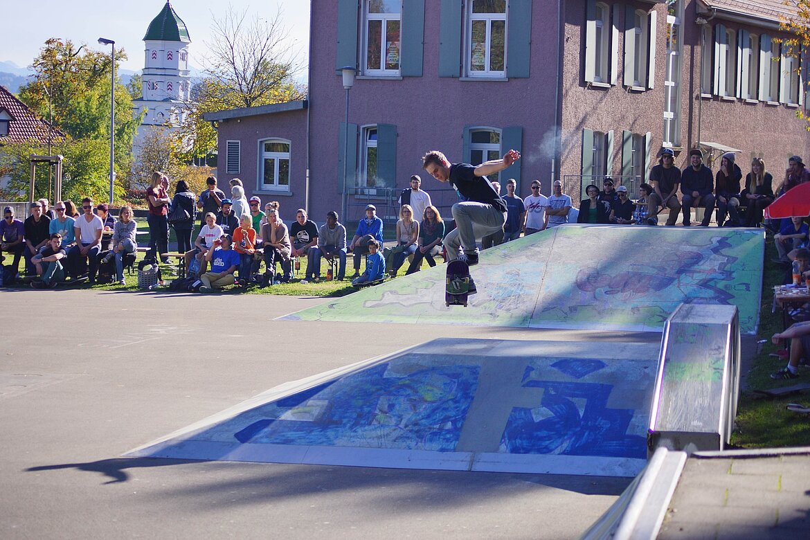 Ein Skateboardfahrer springt über eine Rampe, während eine große Menge von Zuschauern auf einer Wiese sitzt. Im Hintergrund sind Gebäude und Bäume sichtbar, sowie ein Turm. Der Himmel ist klar und sonnig.
