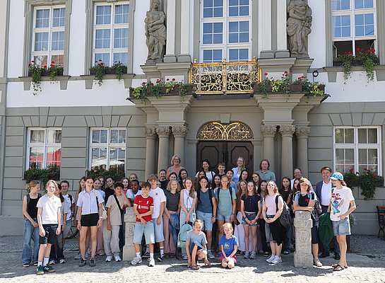 Eine große Gruppe von Schülerinnen und Schülern steht vor einem historischen Gebäude mit einer dekorativen Fassade. Einige Personen lächeln in die Kamera. Blumenkästen schmücken die Fenster, und die Atmosphäre wirkt fröhlich und einladend.