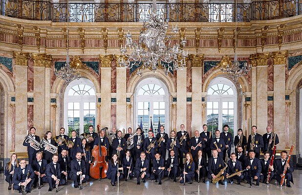 Eine große Gruppe von Musikern, die in formeller Kleidung, hauptsächlich Schwarz, posieren. Sie stehen in einem prunkvollen Raum mit hohen Fenstern und goldenen Verzierungen. Verschiedene Instrumente wie Trompeten, Klarinetten und ein Kontrabass sind zu sehen.