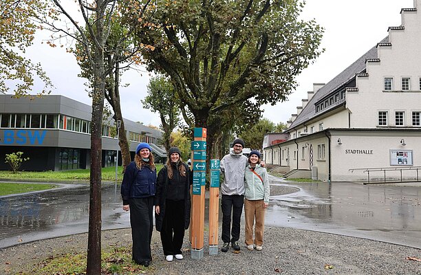 Eine Gruppe von vier Personen steht an einem Wegweiser in einer städtischen Umgebung. Im Hintergrund sind Gebäude und ein Baum zu sehen. Der Boden ist nass, vermutlich nach Regen. Die Personen tragen verschiedene Regenjacken und haben lässige Kleidung an.