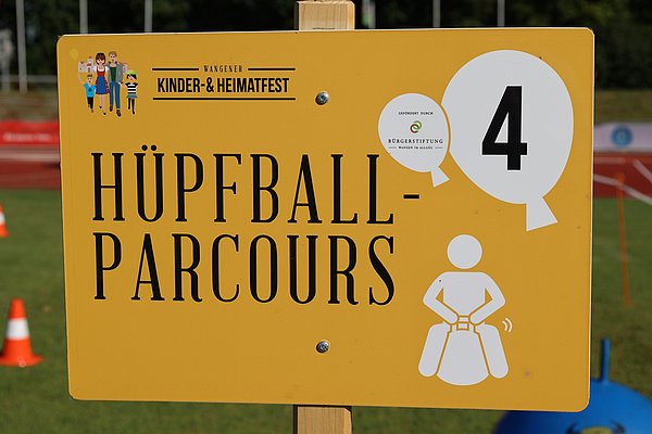 Ein gelbes Schild mit der Aufschrift "Hüpfball-Parcours" und der Nummer 4. Daneben ist ein Symbol einer Person, die auf einem Hüpfball sitzt. Im oberen Bereich sind Figuren einer Familie abgebildet. Im Hintergrund sind weitere Sportgeräte und Verkehrshütchen sichtbar.