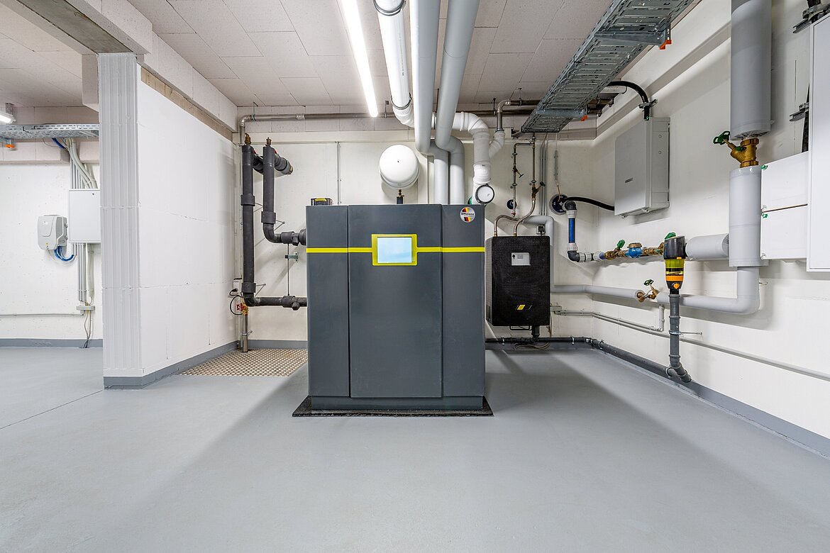 Ein Technikraum mit einem großen, modernen Heizungsgerät in der Mitte. Rohrleitungen verlaufen an den Wänden und Decke. Der Boden ist grau und gut beleuchtet.