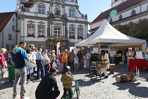 Auf einem Platz stehen viele Menschen vor einem Festzelt, während ein Musiker mit Percussion einen Rhythmus spielt. Im Hintergrund ist ein historisches Gebäude zu sehen, während einige Kinder mit einem Fahrrad spielen. Die Atmosphäre ist lebhaft und festlich.