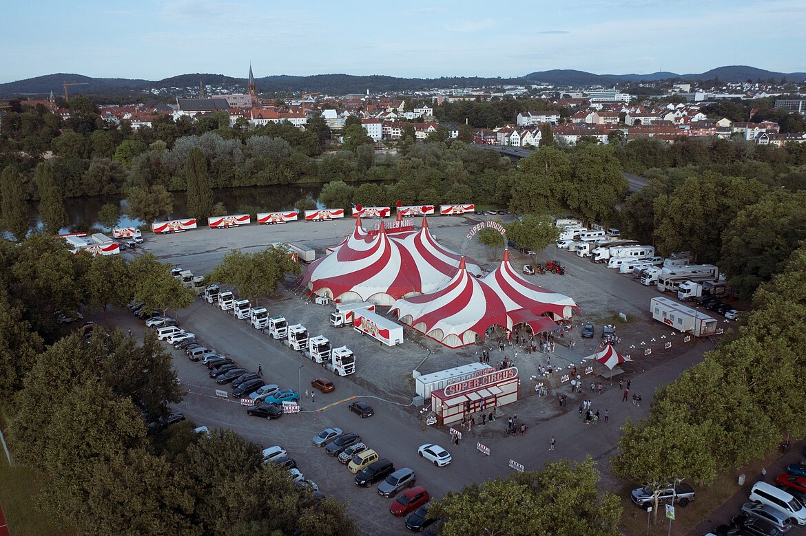 Luftaufnahme eines Zirkusgeländes mit rot-weißen Zelten und vielen Fahrzeugen auf einem Parkplatz. Umgeben von Bäumen und einer hügeligen Landschaft. Im Hintergrund sind Wohnwagen und eine Gruppe von Menschen zu sehen.