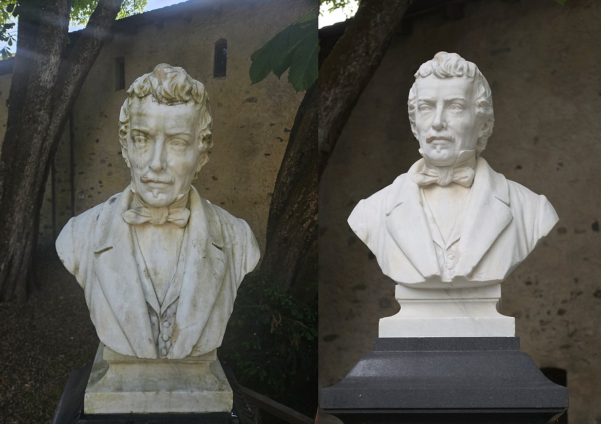 Eine Marmorbüste eines Mannes mit lockigem Haar, Anzug und Fliege, steht auf einem Sockel. Die Statue wird von Bäumen umgeben und hat eine natürliche Hintergrundszene. Sie zeigt einen nachdenklichen Ausdruck und ist sorgfältig bearbeitet.