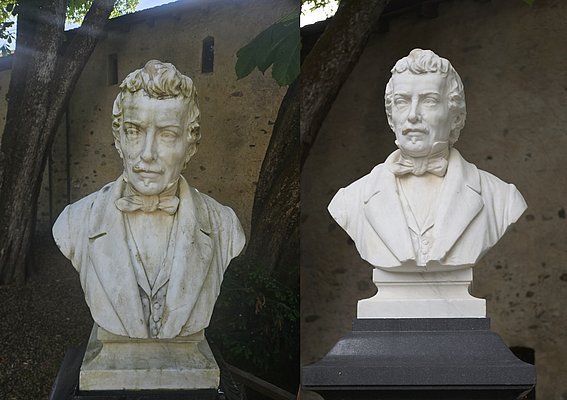 Eine Marmorbüste eines Mannes mit lockigem Haar, Anzug und Fliege, steht auf einem Sockel. Die Statue wird von Bäumen umgeben und hat eine natürliche Hintergrundszene. Sie zeigt einen nachdenklichen Ausdruck und ist sorgfältig bearbeitet.