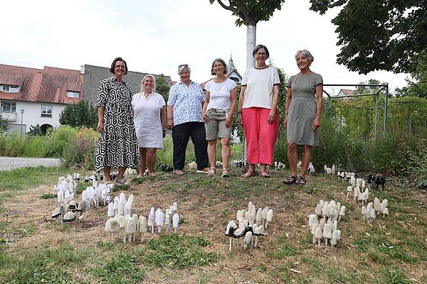 Sechs Frauen stehen auf einem kleinen Hügel in einem Garten, umgeben von einer Vielzahl von kleinen, weißen Figuren, die wie Pilze aussehen. Sie lächeln und posieren für das Foto, während im Hintergrund einige Gebäude und Bäume sichtbar sind.