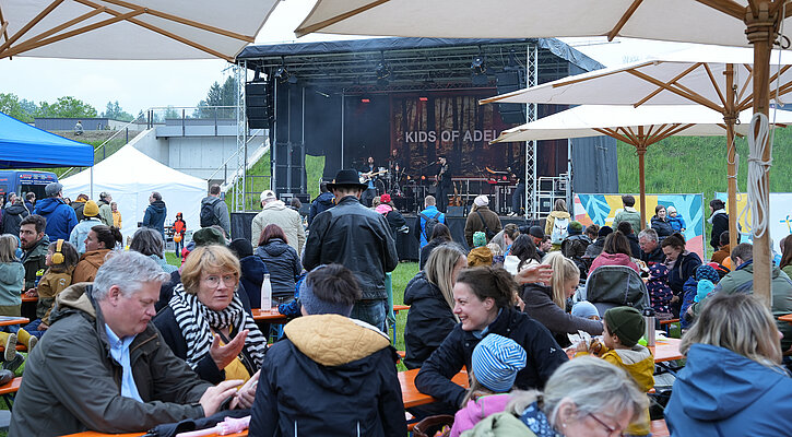 Menschen sitzen an Biertischen vor der Bühne auf der Festwiese