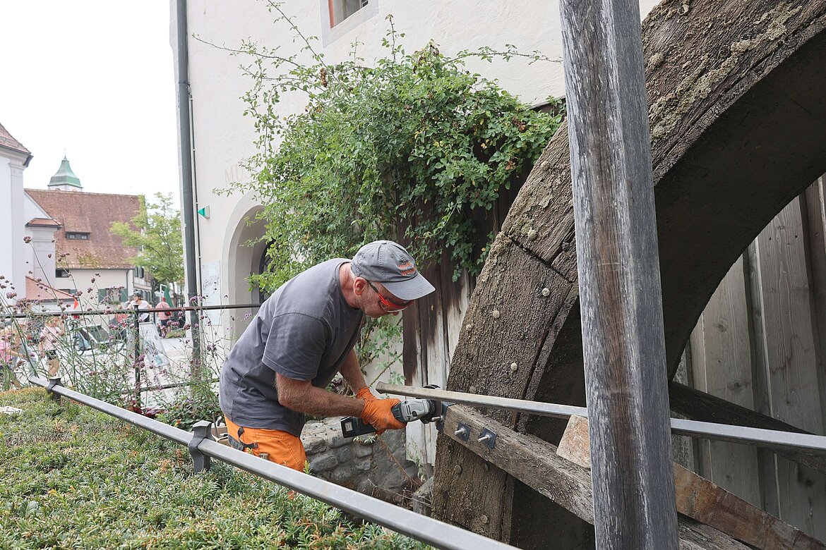 Ein Handwerker arbeitet an einem großen Wasserrad, während er ein Werkzeug in der Hand hält. Er trägt eine graue T-Shirt und orangefarbene Hose, steht in einer natürlichen Umgebung mit Pflanzen und einem historischen Gebäude im Hintergrund.