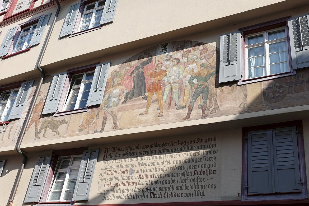 Das Bild zeigt eine Wand mit einem historischen Wandgemälde, das eine Szene mit mehreren Personen in historischen Kostümen darstellt. Einige Fenster mit blauen Fensterläden sind sichtbar, und darunter befindet sich ein Text in altdeutscher Schrift.