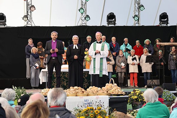 Zwei Pfarrer und eine Pfarrerin, dahinter ein Chor auf großer Bühne