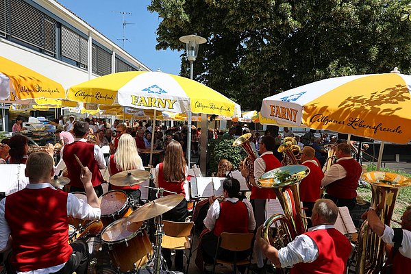 Musikkapelle vor einem Platz voller Menschen unter Sonnenschirmen.