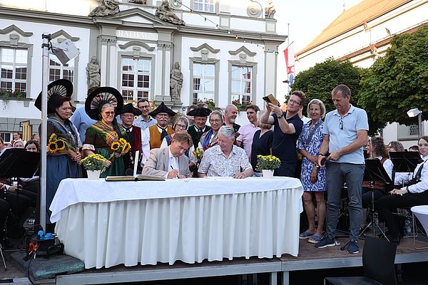 Eine Gruppe von Menschen steht um einen Tisch, an dem zwei Männer ein Dokument unterzeichnen. Im Hintergrund sind Ornamente und Statuen an einem Gebäude zu sehen, während einige Personen Trachten tragen und Sonnenblumen halten. Es herrscht festliche Stimmung.