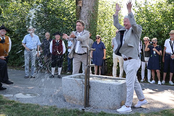 Zwei Männer in Anzügen stehen an einem Brunnen, aus dem Wasser spritzt. Sie heben die Hände in die Luft und lachen. Im Hintergrund sind eine Gruppe von Zuschauern in festlicher Kleidung zu sehen, die zuschauen.