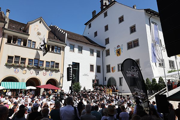 Ein belebter Platz mit mehreren Menschen, die einem Konzert oder einer Veranstaltung zuhören. Im Hintergrund sind historische Gebäude mit bunten Wappen und Fahnen zu sehen, darunter ein Kranz von blühenden Bäumen. Der Himmel ist blau und sonnig.
