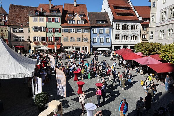 Ein belebter Marktplatz mit bunten Gebäuden, Tischen und Sonnenschirmen. Menschen unterschiedlicher Altersgruppen stehen und interagieren, während ein Zelt im Vordergrund sichtbar ist. Es ist sonnig und festliche Stimmung herrscht.
