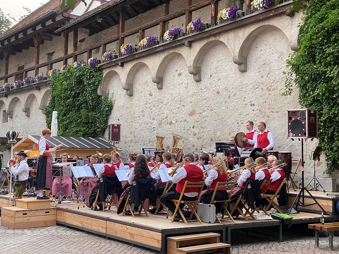 Eine Blasmusikkapelle tritt auf einer Bühne in einem Innenhof auf. Die Musiker tragen traditionelle Kleidung mit roten Westen. Im Hintergrund sind Blumen und eine historische Steinwand sichtbar. Der Dirigent leitet die Musiker, während verschiedene Instrumente gespielt werden.