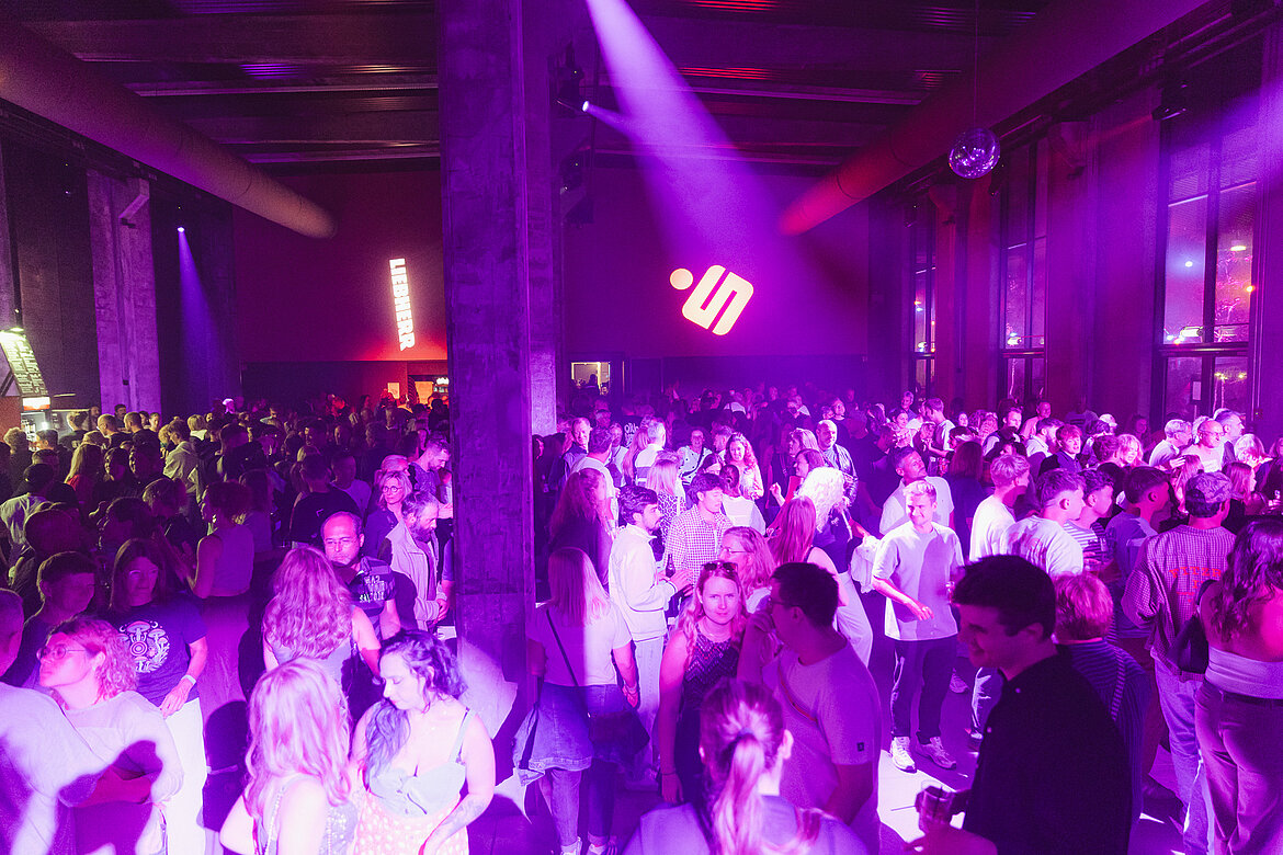 Eine lebhafte Partyszene mit vielen Tänzern in einem Raum, beleuchtet von bunten Lichtern. Menschen verschiedener Altersgruppen interagieren und tanzen in einer geselligen Atmosphäre. Im Hintergrund sind dunkle Wände und große Fenster zu sehen.