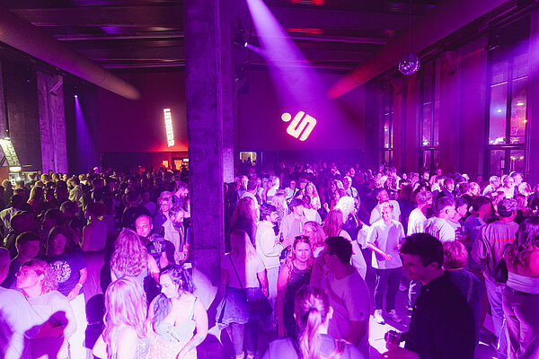 Eine lebhafte Partyszene mit vielen Tänzern in einem Raum, beleuchtet von bunten Lichtern. Menschen verschiedener Altersgruppen interagieren und tanzen in einer geselligen Atmosphäre. Im Hintergrund sind dunkle Wände und große Fenster zu sehen.