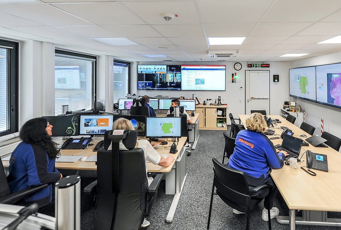 Ein Raum mit mehreren Arbeitsplätzen, an denen Mitarbeiter vor Computerbildschirmen sitzen. Es gibt große Wandbildschirme mit Karten und Daten. Die Atmosphäre wirkt geschäftig und organisiert, während die Mitarbeiter konzentriert arbeiten.