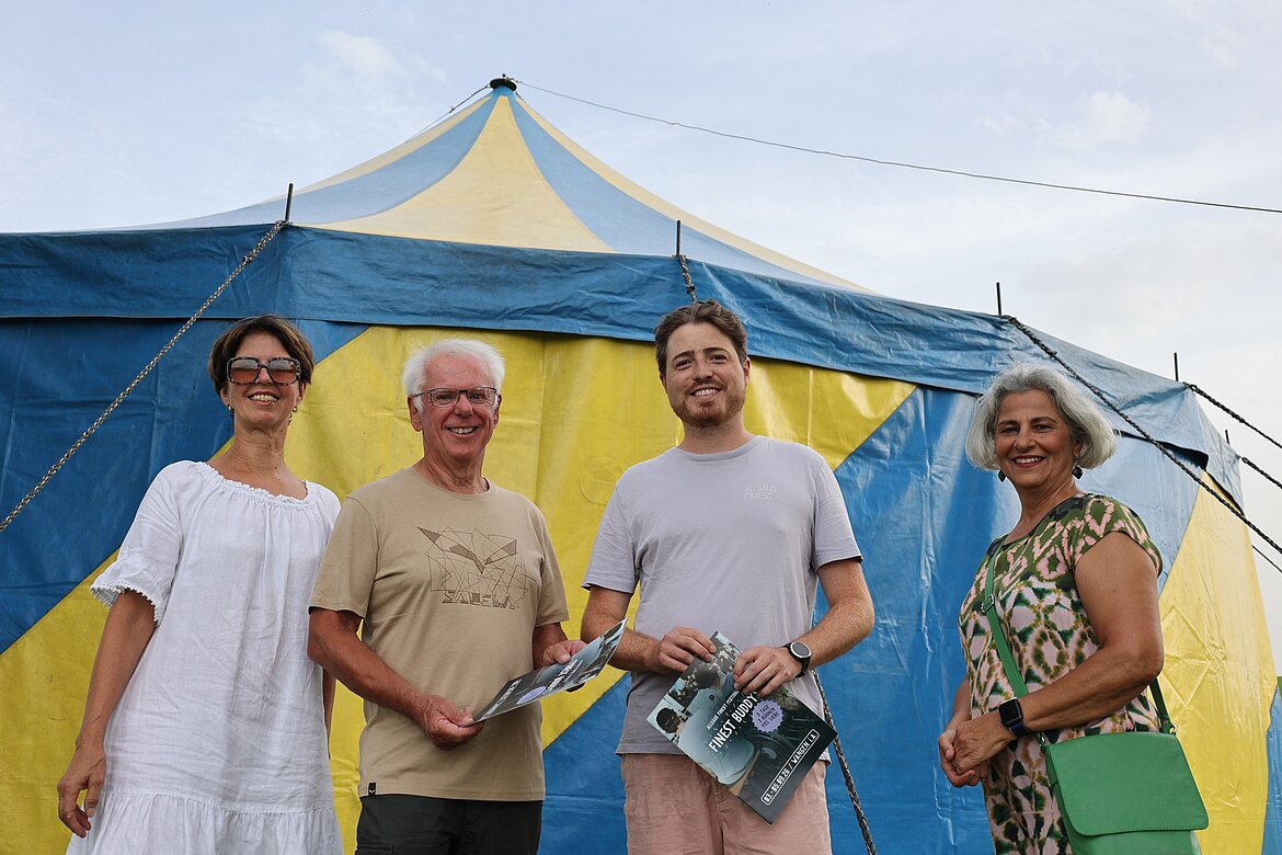 Eine Gruppe von vier Personen steht vor einem bunten Zelt. Sie lächeln und halten Broschüren in den Händen. Die Personen tragen verschiedene Outfits: eine weiße Bluse, ein beige T-Shirt, ein graues T-Shirt und ein gemustertes Kleid. Der Himmel ist teilweise bewölkt.