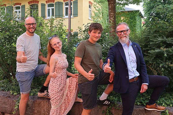 Vier Personen posieren lächelnd im Freien. Sie zeigen Daumen nach oben und stehen vor einer grünen Hecke und einem gelben Gebäude im Hintergrund. Die Gruppe besteht aus einem Mann in Anzug, einem weiteren Mann in Freizeitkleidung und zwei Jugendlichen.
