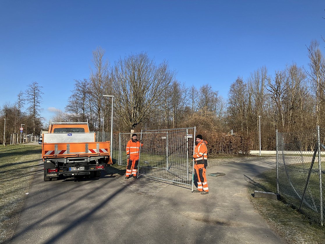 Zwei Männer vom Bauhof heben einen Bauzaun aus der Verankerung. Daneben steht ein Transportfahrzeug. 