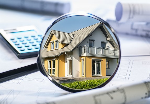 Ein vergrößertes Bild eines Hauses, das in einer Lupe betrachtet wird. Das Haus hat ein gelbes Äußeres mit einem Satteldach und einem Balkon. Im Hintergrund sind Baupläne und ein Taschenrechner sichtbar.