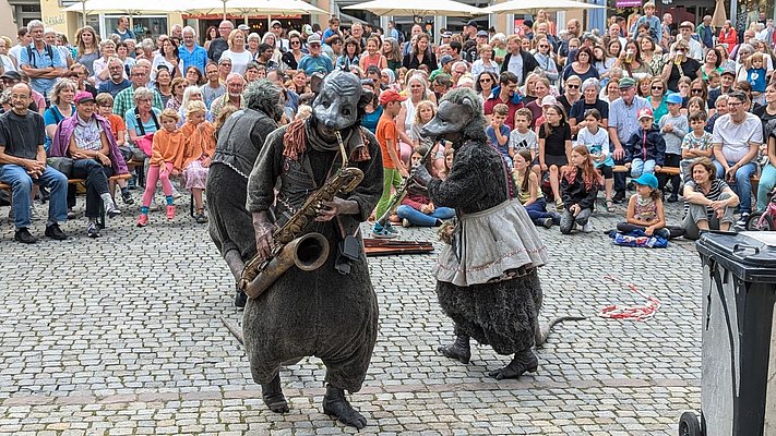 Eine Gruppe von Menschen in einem Platz beobachtet eine Straßenperformance, bei der zwei Akteure in Rattenkostümen spielen. Einer spielt ein Saxophon, während der andere eine Art Tanz ausführt. Die Zuschauer, darunter viele Kinder, sind fasziniert und applaudieren.