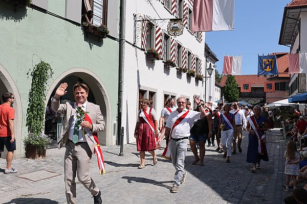 Eine Feierlichkeit in einer Stadtstraße, wo Menschen in festlicher Kleidung mit Bändern und Blumenparade. Einige Teilnehmer winken der Menge zu. Die Umgebung zeigt historische Gebäude und eine festliche Dekoration mit Fahnen.