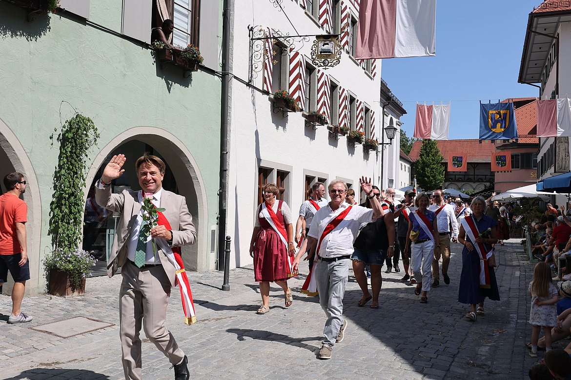 Eine Feierlichkeit in einer Stadtstraße, wo Menschen in festlicher Kleidung mit Bändern und Blumenparade. Einige Teilnehmer winken der Menge zu. Die Umgebung zeigt historische Gebäude und eine festliche Dekoration mit Fahnen.
