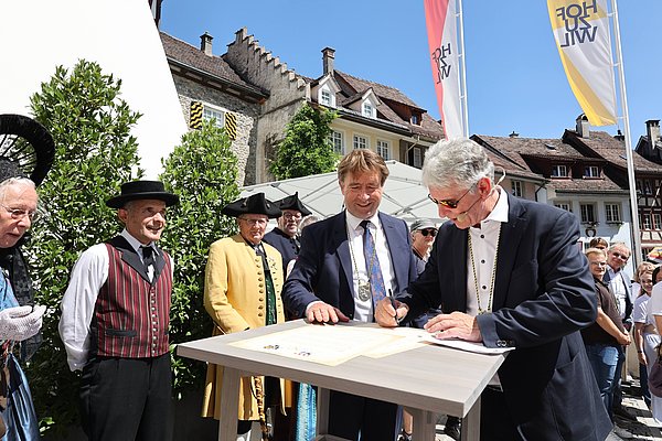 Eine Gruppe von Personen in traditioneller Kleidung steht um einen Tisch, auf dem eine Urkunde liegt. Zwei Männer, einer mit Brille, unterschreiben das Dokument. Im Hintergrund sieht man Zuschauer und historische Gebäude bei sonnigem Wetter.