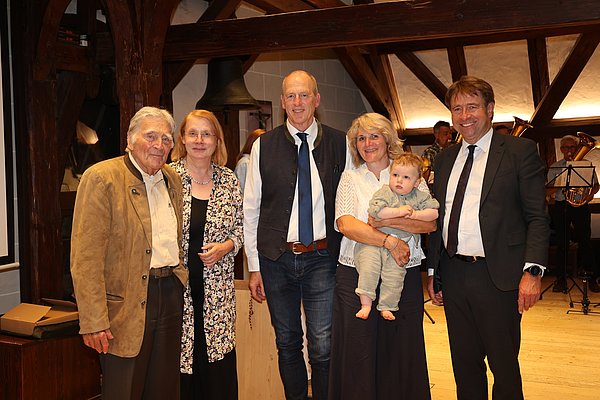 Eine Gruppe von fünf Personen steht in einem Raum mit Holzbalken. Es sind drei Erwachsene und ein Baby zu sehen. Die Menschen lächeln und posieren für das Foto, im Hintergrund sind unscharfe Musiker zu erkennen.
