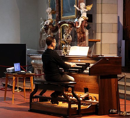 Ein Organist sitzt an einer Kirchenorgel und spielt. Er trägt eine Brille und hat einen Laptop neben sich auf einem Tisch. Im Hintergrund sind historische Kunstwerke und Statuen zu sehen. Der Raum hat eine kirchliche Atmosphäre.