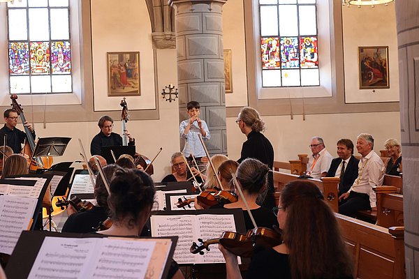 Ein Orchester spielt in einem Kirchensaal mit bunten Fenstern. Musiker auf der Bühne und im Publikum sind gut zu sehen. Eine Dirigentin leitet das Ensemble, während einige Zuhörer entspannt auf der Bank sitzen. Die Atmosphäre ist festlich und konzentriert.