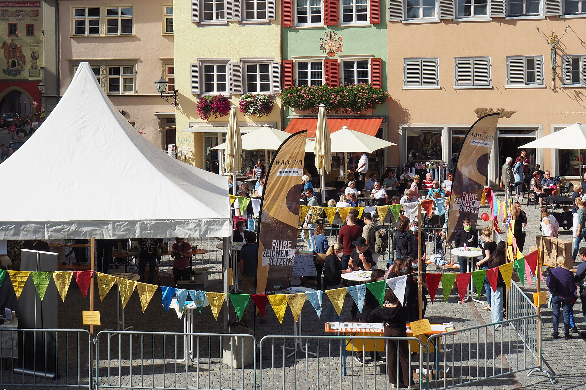Markt mit Festzelten und bunten Fahnen, umgeben von Menschen, die an Tischen sitzen oder stehen. Im Hintergrund historische Gebäude mit Fensterläden und Blumenverzierungen. Sonniges Wetter und eine lebhafte Atmosphäre.