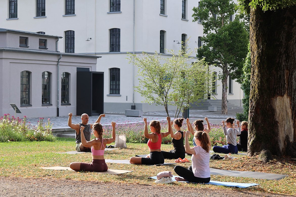 Gruppe von Menschen, die im Freien Yoga praktizieren. Sie sitzen auf Matten und heben ihre Arme, während sie von einer modernen Gebäudeumgebung umgeben sind. Bäume und Blumen sind im Hintergrund zu sehen.