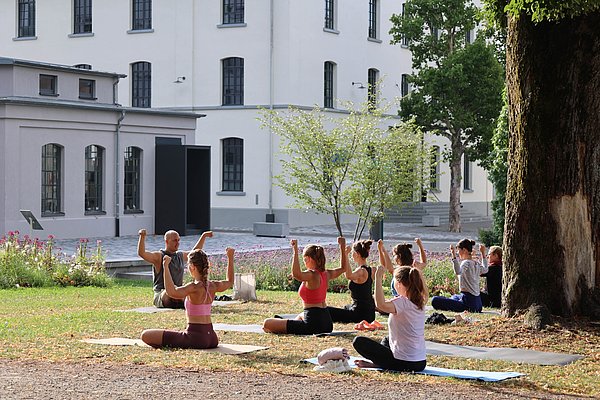 Gruppe von Menschen, die im Freien Yoga praktizieren. Sie sitzen auf Matten und heben ihre Arme, während sie von einer modernen Gebäudeumgebung umgeben sind. Bäume und Blumen sind im Hintergrund zu sehen.