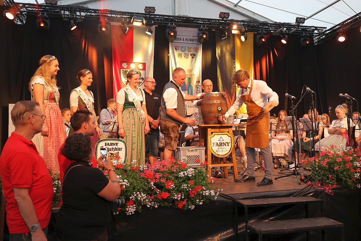 Auf einer Bühne im Festzelt steht ein Bierfass, in das ein Hahn geschlagen wird