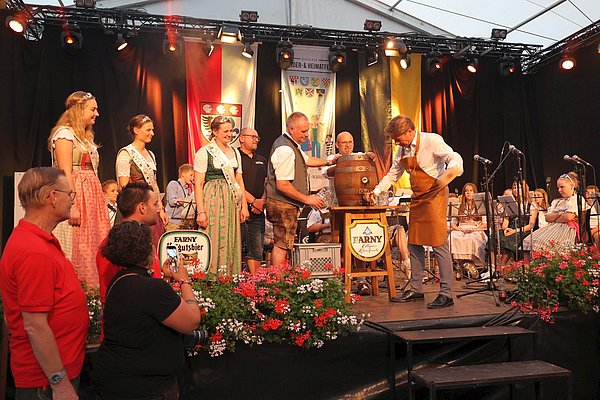 Auf einer Bühne im Festzelt steht ein Bierfass, in das ein Hahn geschlagen wird