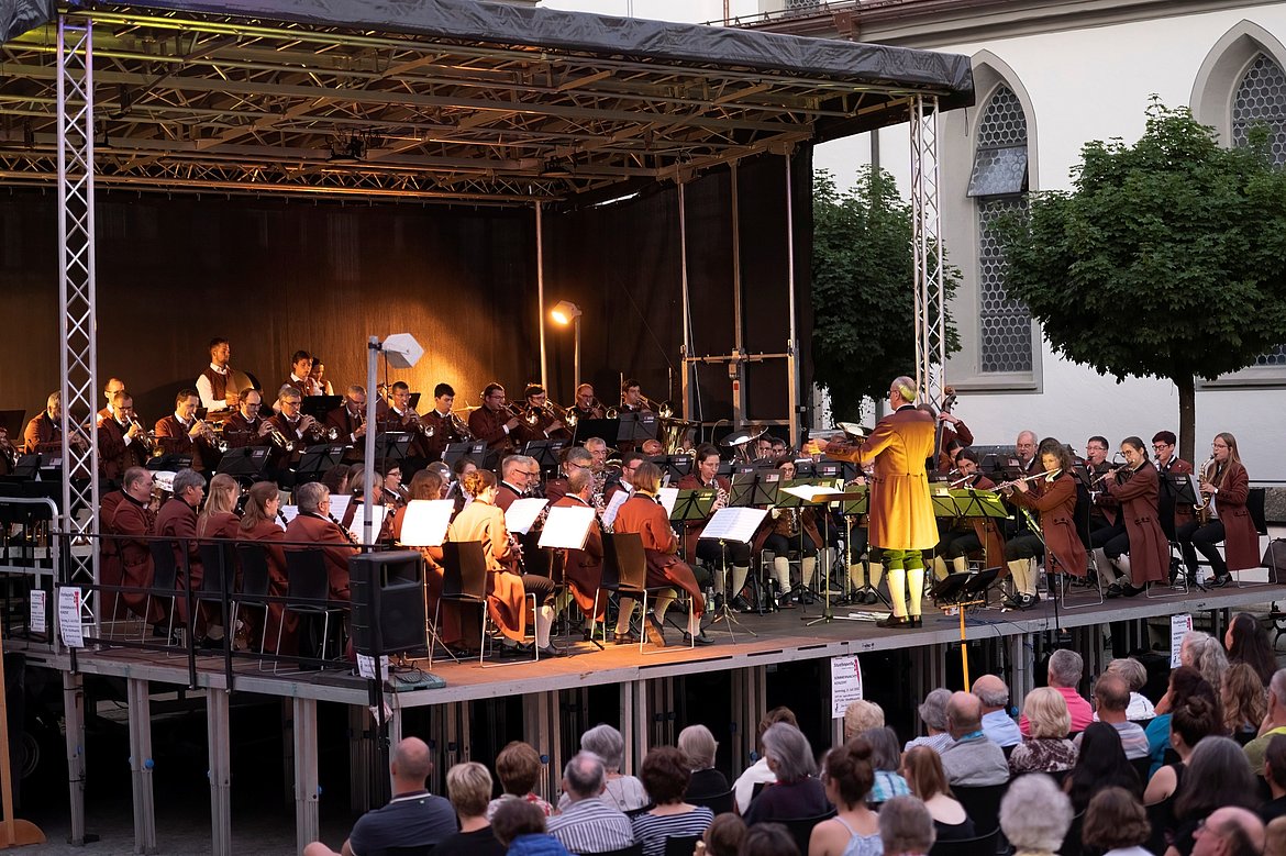 Ein Orchester spielt auf einer Freiluftbühne vor Publikum. Der Dirigent steht vorne und leitet die Musiker, die in historischen Kostümen gekleidet sind. Im Hintergrund sind Bäume und ein Gebäude zu sehen. Zuschauer sitzen in Reihen und hören gebannt zu.