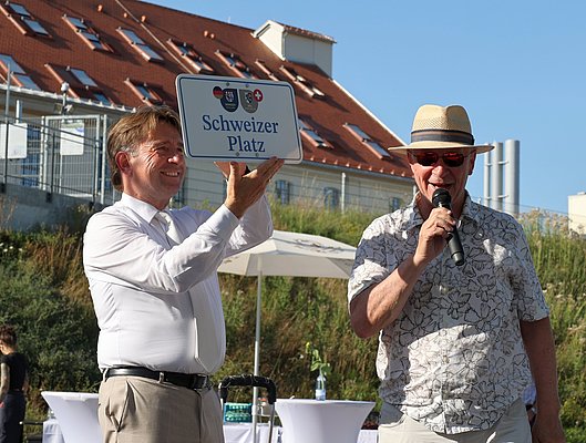 Zwei Männer stehen im Freien bei einer Veranstaltung. Der Mann links hält ein Schild mit der Aufschrift "Schweizer Platz" hoch, während der Mann rechts ins Mikrofon spricht. Im Hintergrund sind Zelte und die Skyline zu sehen.
