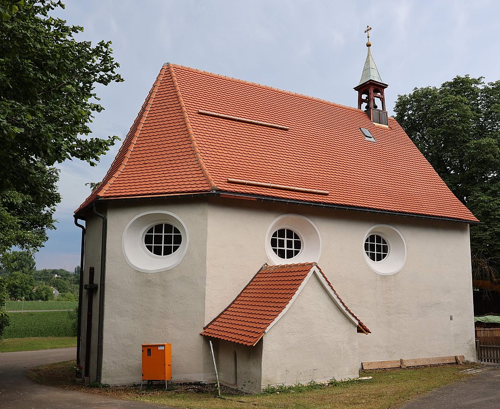 Ein kleines, hellgraues Gebäude mit einem rot gedeckten, spitzen Dach und einem hohen, turmartigen Aufsatz. Die Wände haben runde Fenster. Vor dem Gebäude steht eine orangefarbene Box. Im Hintergrund ist eine grüne Wiese mit Bäumen sichtbar.