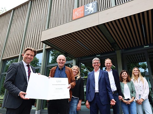 Zwei Männer halten eine große Urkunde in der Hand. Weiter Personen stehen daneben.